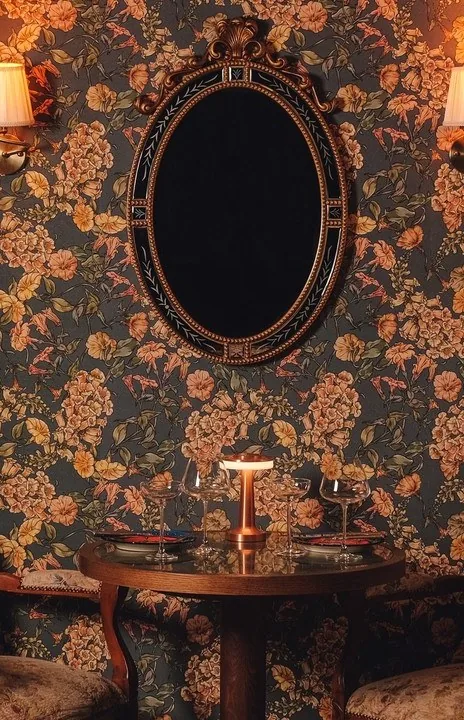 An oval mirror on floral damask wallpaper above a small table lamp
