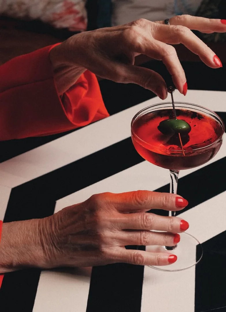 A red cocktail with an olive, held over a black-and-white striped table